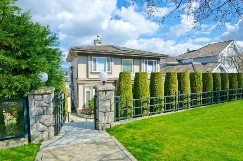 Big opulent home behind nicely trimmed trees with turquoise iron fence in the suburbs