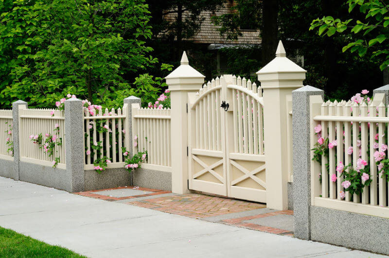 residential house with fencing and gate made from wood, brick, and granite, pink rose bushes come through picket posts